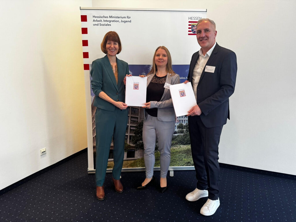 Die hessische Ministerin für Arbeit, Integration, Jugend und Soziales Heike Hofmann, Marion Lepold von der QiK Onlineakademie und Prof. Dr. Timm Albers von der Universität Paderborn stehen vor einem Roll-Up mit dem Hessener Logo und halten stolz Mappen, auch mit dem Hessen-Logo darauf, in die Kamera.
