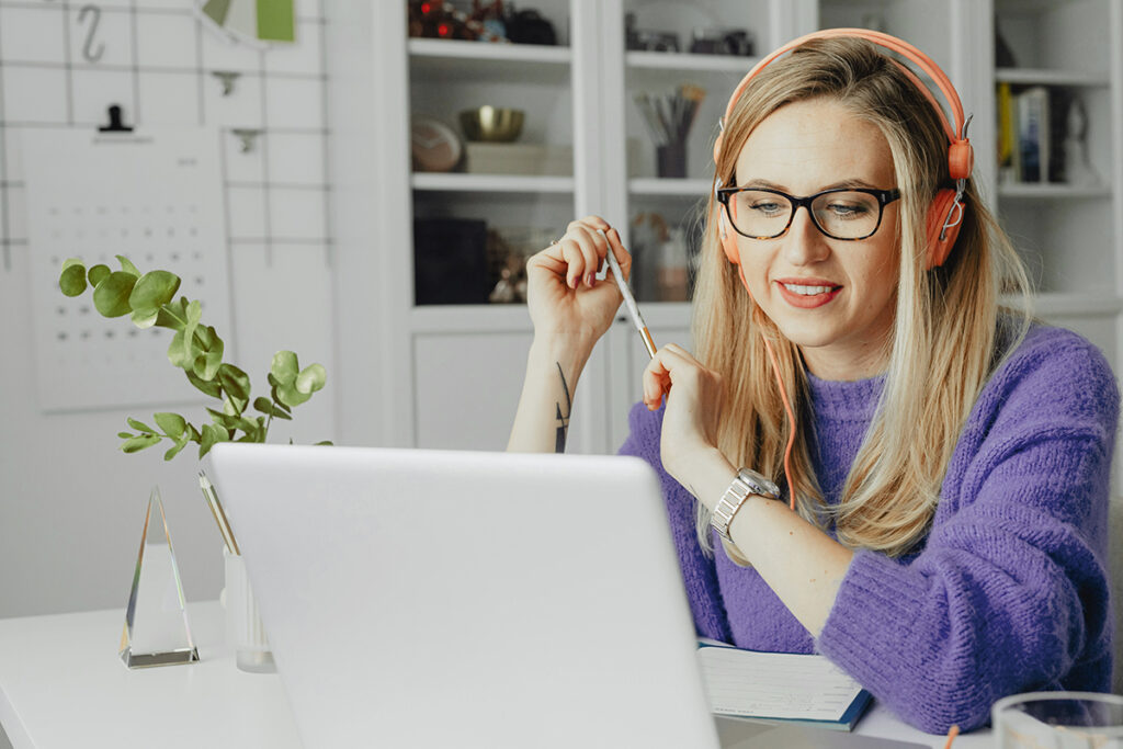 Eine Frau sitzt in einem Arbeitszimmer am Schreibtisch und schaut auf ihren Laptop. Sie trägt einen violetten Pullover und hat Kopfhörer auf.