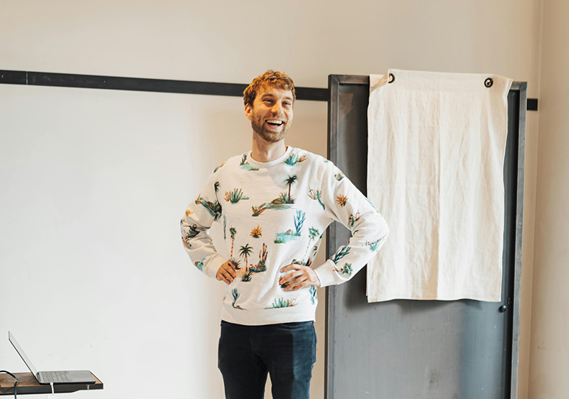 Ein Mann in einem bunten Pulli steht lachend und mit den Händen auf seine Hüften gestützt vor einer Wand und hält einen Vortrag.