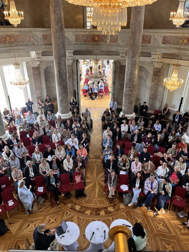 Ein prunkvoller Saal im Schloss Bellevue, gefüllt mit Sitzen und Publikum, Perspektive von der Empore blickend und der Bühne im unteren Bildbereich.