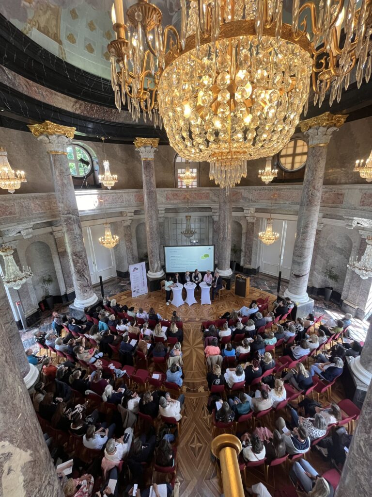 Ein prunkvoller Saal im Schloss Bellevue, gefüllt mit Sitzen und Publikum, vorne ein Bühnenbereich, Perspektive von der Empore blickend mit einem Kronleuchter oben im Blickfeld.