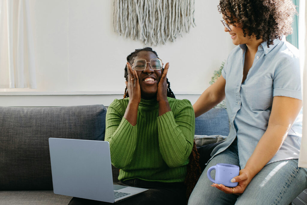 Zwei Frauen sitzen in einem hellen Zimmer auf einer Couch. Eine hat einen Laptop auf dem Schoß und hebt in Freude ihre Hände an ihre Wangen. Die zweite Frau schaut sie an und lacht freundlich.